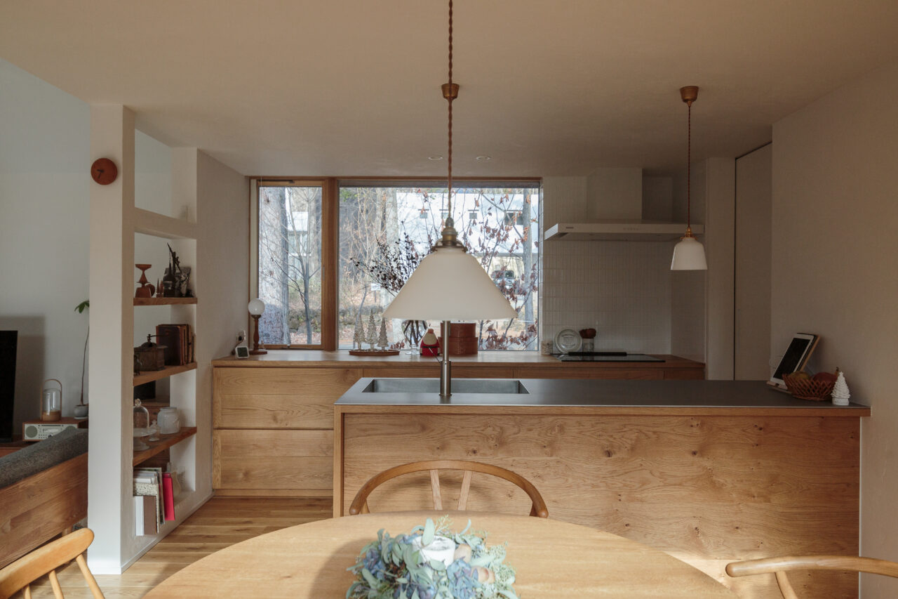 造作の木製キッチンとアイランドカウンター、無垢材の収納と大きな窓から自然光が差し込む温かみのあるダイニング空間